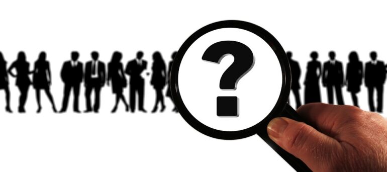 A magnifying glass with a questionmark over black silhouettes of people. 